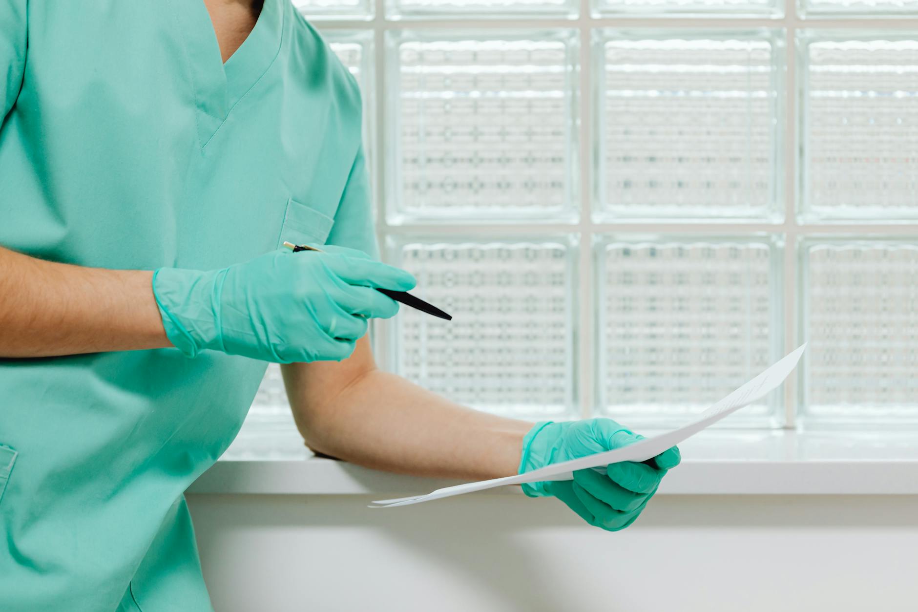 A healthcare worker in scrubs and gloves holding a pen and paper in a medical facility.
