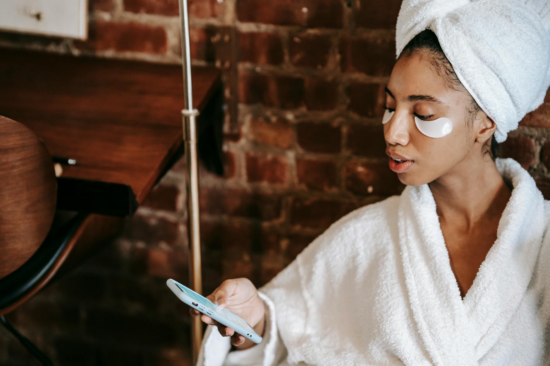 Young ethnic female with eye patches surfing internet on cellphone during facial procedure in spa center
