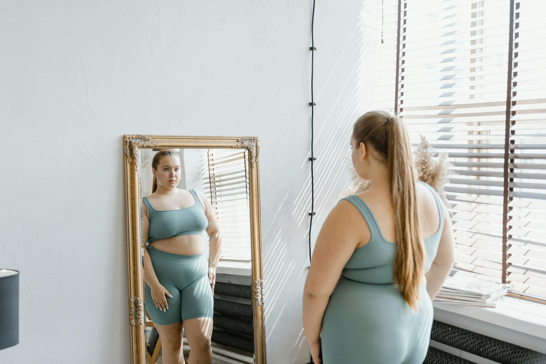 Woman in activewear embraces body positivity, standing before a mirror indoors.