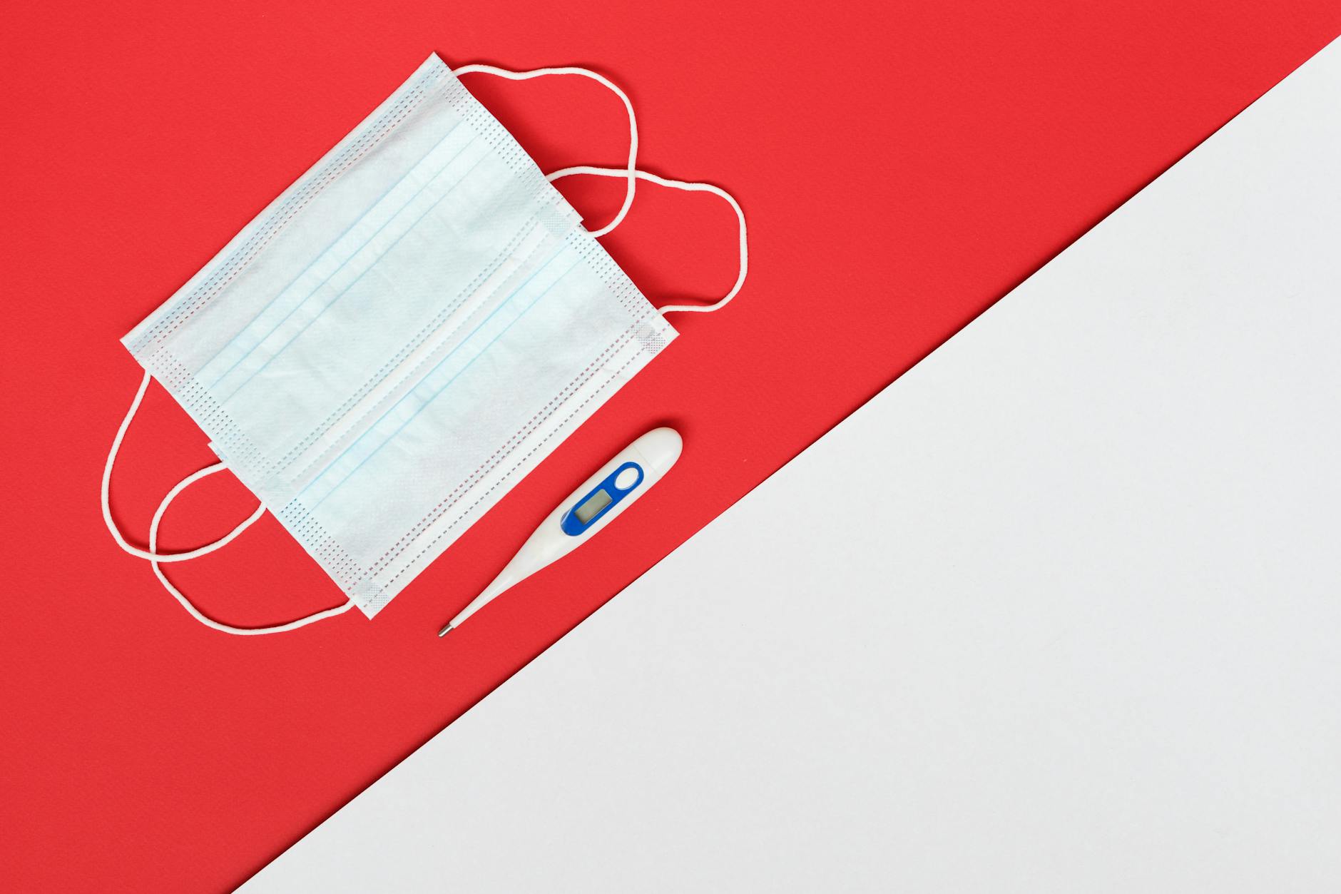 Flat lay of a face mask and digital thermometer on red and white background.