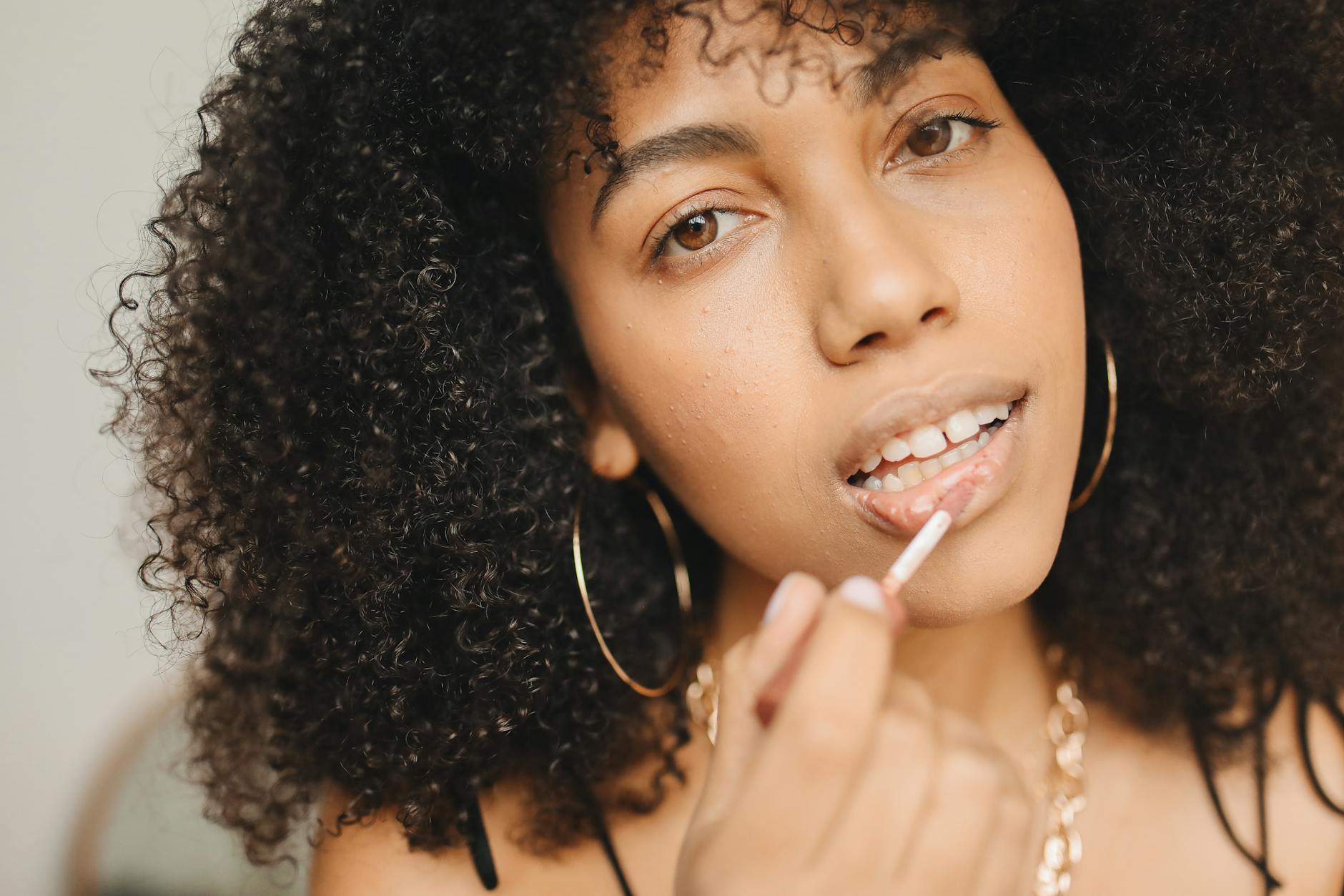 Close-up of a black woman with curly hair applying lip gloss, preparing for an event indoors.