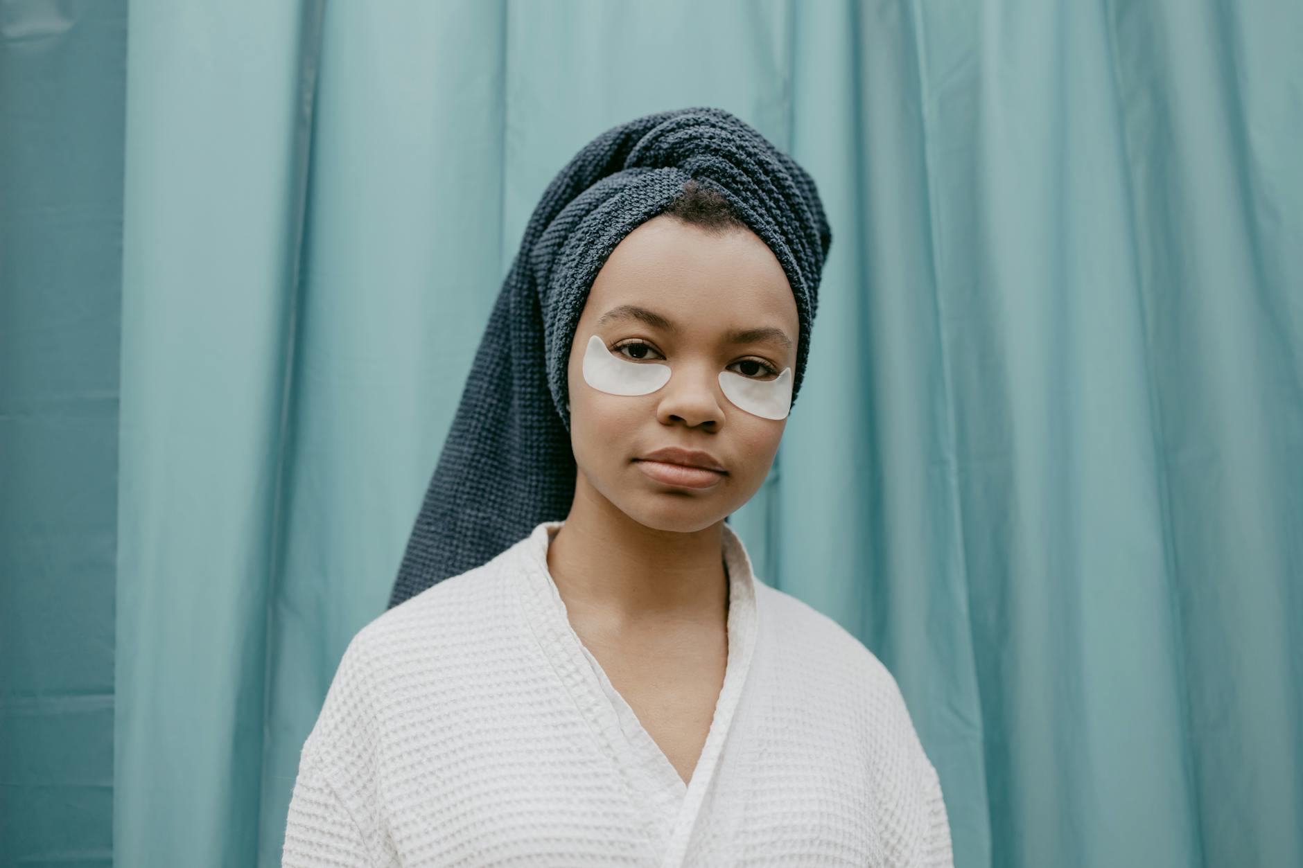 Woman in a white robe wearing eye patches, standing indoors with a head towel against a curtain backdrop.